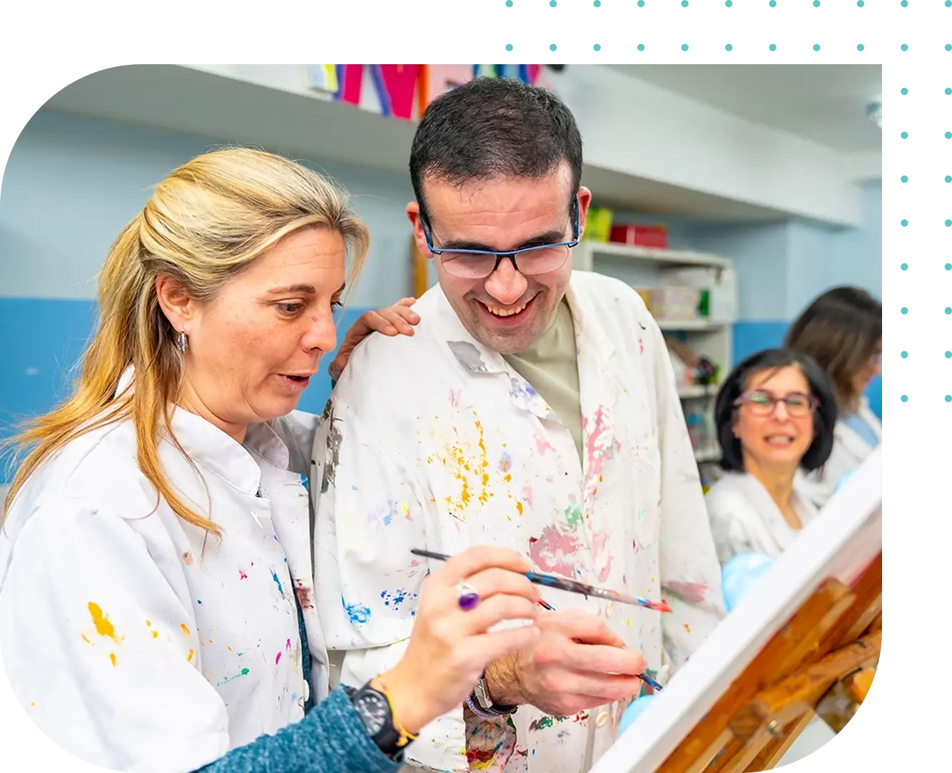 Two people wearing paint-splattered lab coats enjoy painting in a classroom, supported by Disabilities Support Services, while a third person sits and observes in the background.